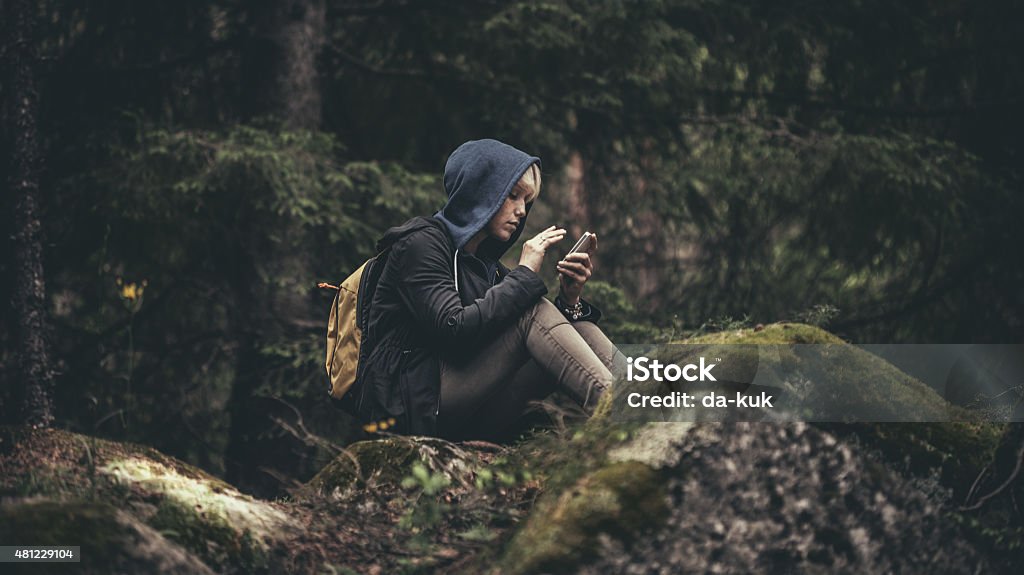 Viaggiatore con zaino seduto nella foresta e con un smartphone - Foto stock royalty-free di Telefono cellulare