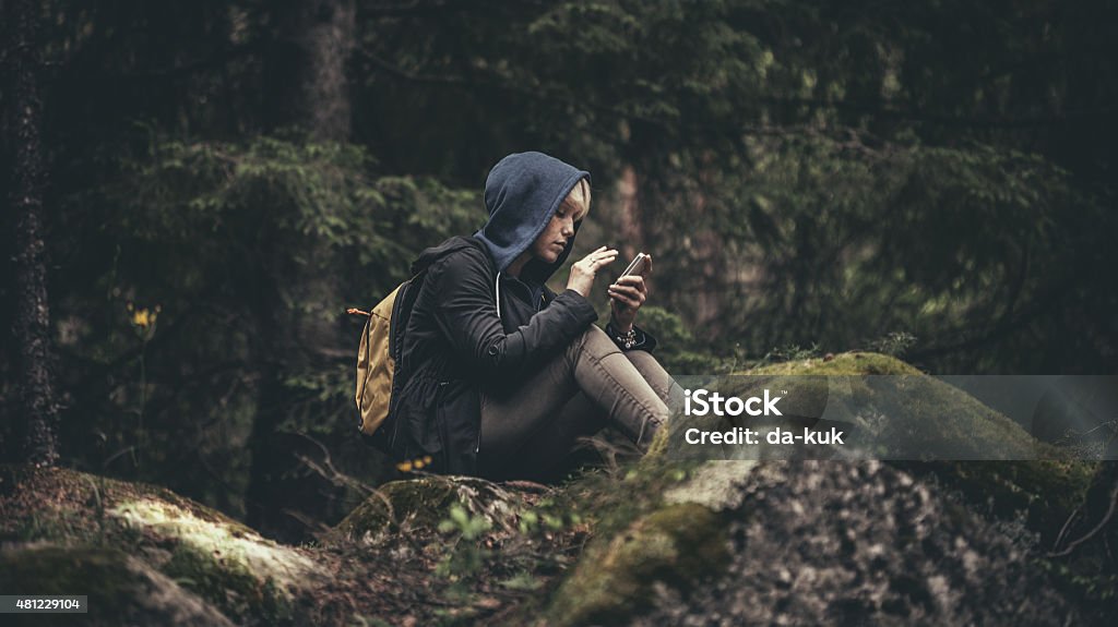 Viajante de mochila sentado na floresta e segurando um smartphone - Foto de stock de Telefone celular royalty-free