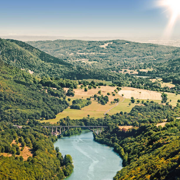 Square composition photography of countryside landscape of Ain river valley, with tree forest and arch bridge crossing the preserved natural Ain river. Image taken in Bugey, Rhone-Alpes region in France (Europe). This picture was made during a bright beautiful sunset on field. Near to Alps chain and Jura in Franche Comte. Shot in middle of summer season with colorful tree and hill, with an impressive unobstructed view on rolling landscape mountain under sunlight. It's an aerial view by high angle view.