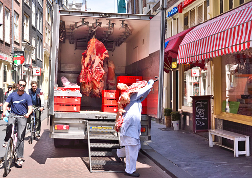 Amsterdam, Netherlands - June 15, 2015: Raw beef, butchery transport. Raw meat hanging on meat hooks in a truck. Man carrying beef into the butcher's shop on a busy street in Amsterdam.