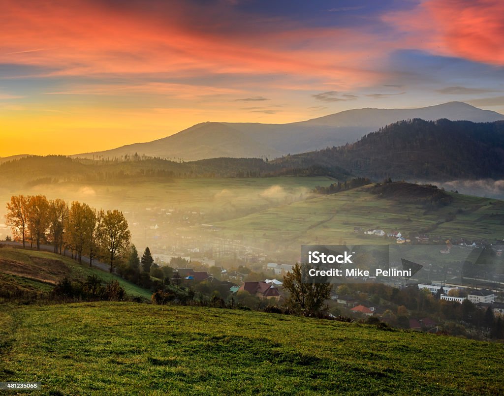 meadow near village in autumn mountains at sunrise village behind the agricultural meadow with yellow trees on hillside in autumn mountains in morning light 2015 Stock Photo