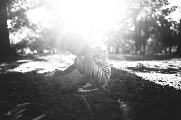 A young, unrecognizable girl plays by herself with sticks and dirt in a park. Natural sunlight shines through the trees with a bright sun flare. A depiction of discovery and learning through sensory contact.  High contrast black and white horizontal image with copy space.
