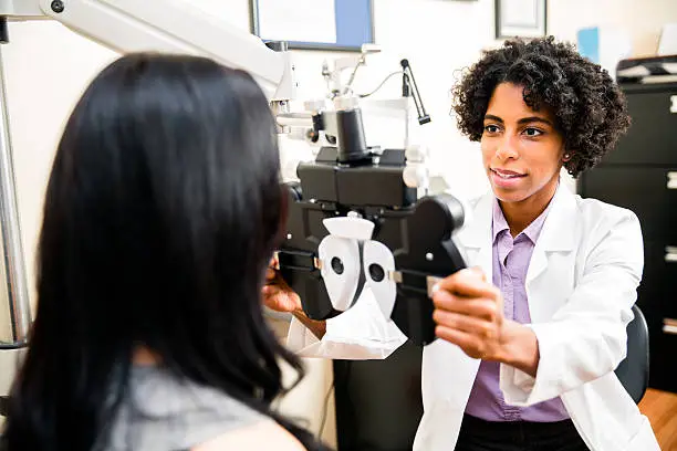 A African American optometrist is checking her patient's eye sight. Optometrist is ethnic of African Decent. Doctor is in ethnic minority. She is checking her patient's eye sight by pulling eye test equipment - Phoropter. Its real optometrist office.    