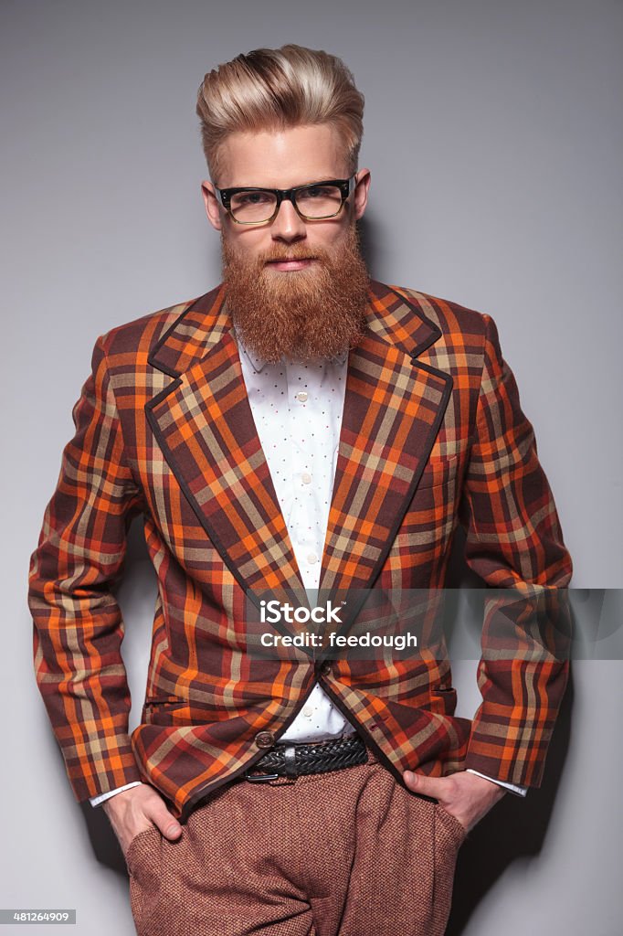 smiling fashion model with long beard leaning against gray wall smiling fashion model with long beard standing with his hands in pockets against a gray studio wall Men Stock Photo