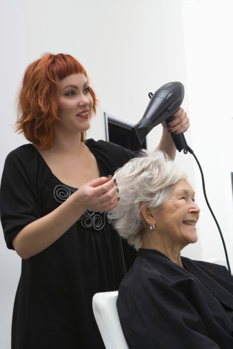 Happy female stylist blow drying senior woman's hair in salon