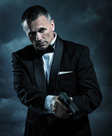 A handsome mature male secret agent, bodyguard, spy, or security staff dressed in an elegant tuxedo and bow tie holding a modern handgun ready to shoot if necessary on a stormy night with cloudy sky in the background.