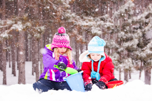 Two young girls (sisters) playing in the snow with toy buckets and shovels.