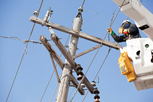 An electric utility lineman is making repairs to a damaged pole. The pole was struck by a car and is leaning, the wires are being removed for the pole's replacement. The text on the yellow bucket bag reads, Always Wear Your Gloves.