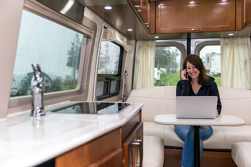 Young Woman Talking on phone and Using a Laptop Inside a luxury Motorhome on a campground on a rainy day.