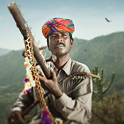 Outdoor image of a folk musician of rural Rajasthan, India playing an ancient bowed violin called Ravanahatha which is a traditional musical instrument. He is in colorful turban and a warm gray shirt playing his music in a beautiful outdoor scene of mountains and sky in a bright day. Square image with selective focus at aperture f/5.6.