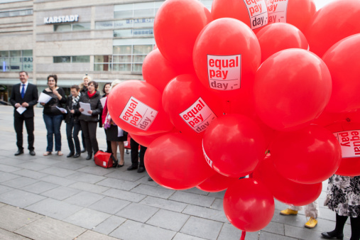 Wiesbaden, Germany - March 21, 2014: Red balloons and participants of Equal Pay Day demonstration in the city center of Wiesbaden. Equal Pay Day is a symbolic day which illustrates how much longer women have to work in a year to earn the same amount of money made by a man in the previous year. The event was organised by BPW - Business and Professional Women Wiesbaden. Some pedestrians in the background