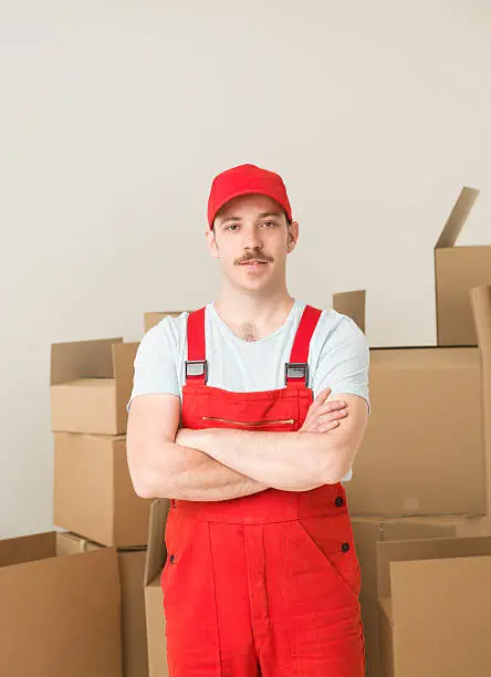 young confident delivery man standing, holding hands crossed against his chest, with cardboard boxes in background