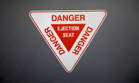 An ejector seat warning sign on the side of a grey painted mlitary aircraft.