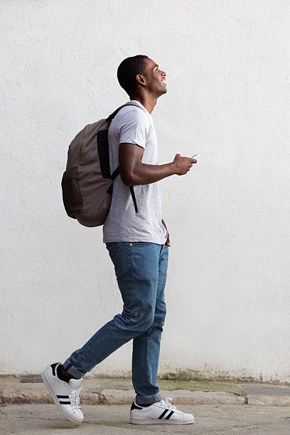 faculdade estudante do sexo masculino sorridente caminhando com mala e telefone celular - tourist backpack smiling cheerful - fotografias e filmes do acervo