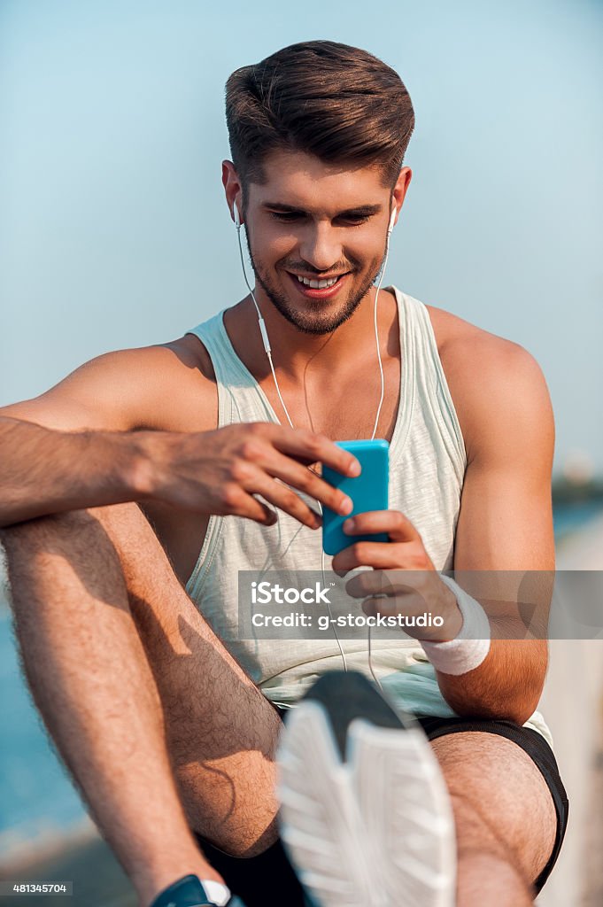 Choosing music for training. Happy young muscular man in headphones looking at this MP3 player and smiling while sitting on parapet 2015 Stock Photo