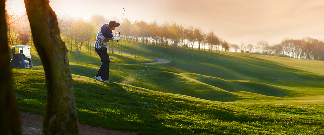 a lone golfer chips onto the green , the low sun is coming from behind him and flaring to camera.