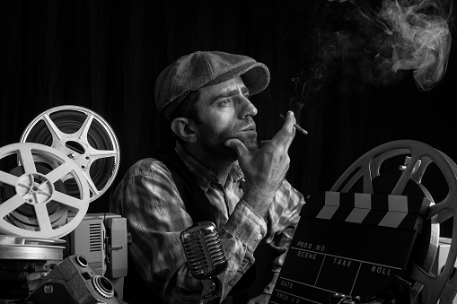 Mid adult man wearing flat cap sitting on desk with film projector, video camera, cinema film reel and clapperboard.He is smoking cigarette in dark.The background is black.Photo is shot with full frame DSLR camera in horizontal composition.