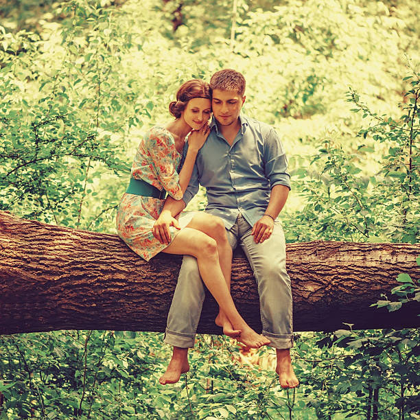 Smiling young loving couple sitting on fallen tree trunk in summer forest
