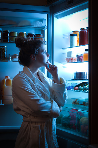 A young caucasian woman standing in front of the open refrigerator at late night, contemplating and wondering about a midnight snack in a domestic home kitchen. She is dressed in a bath robe hungry and looking for food. A symbol of dieting lifestyle. Photographed in vertical format.