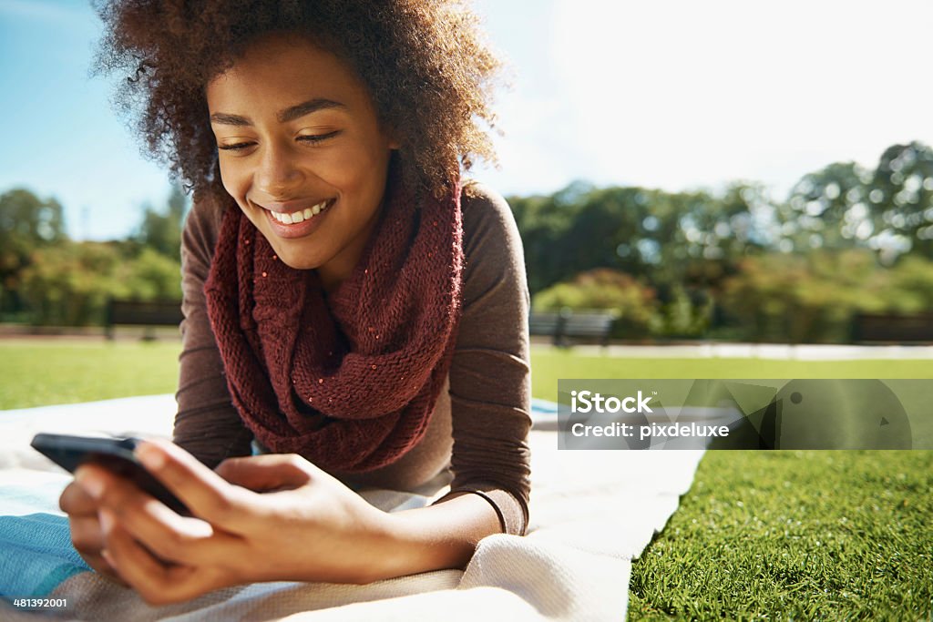 Reading her texts with glee Shot of a smiling young woman relaxing with her phone in the park Lying Down Stock Photo