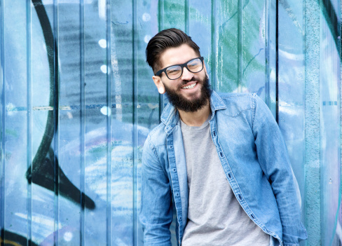Portrait of a young man with beard and glasses laughing outdoors against graffiti background