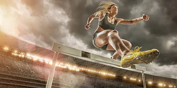 Low angle view of professional athlete jumping over hurdle during race in outdoor athletics stadium full of spectators under dramatic cloudy evening sky at sunset.
