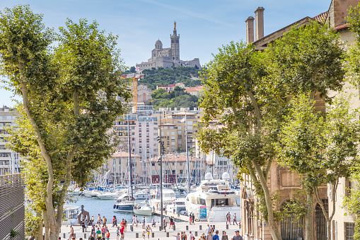 Marseille, France - August 24, 2014: Panoramic view of Basilique Notre-Dame de la Garde from Place Villeneuve-Bargemon near the Vieux Port of Marseille. Many people walking on promenade Quai du Port. Many boats (yachts and sailing boats) at the Pier. In background a yellow crane and the Basilique Notre-Dame de la Garde