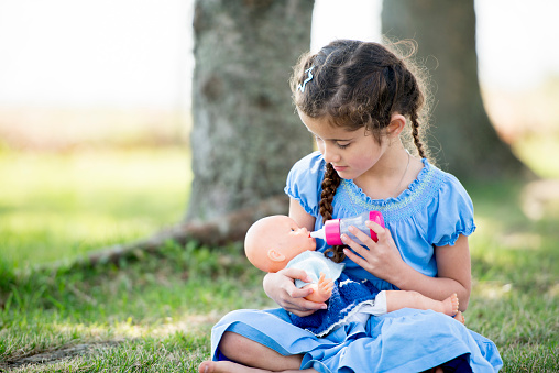 A little girl is sitting outside at the park on the grass, she is holding a doll and pretending to feed the doll with a bottle.