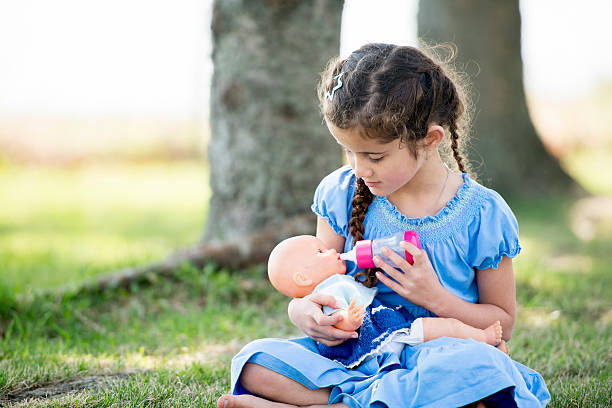 Little Girl Playing with Doll at Park A little girl is sitting outside at the park on the grass, she is holding a doll and pretending to feed the doll with a bottle. girl playing with doll stock pictures, royalty-free photos & images