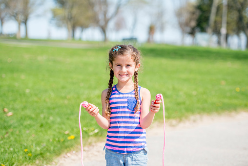 A little girl standing with her jump rope outside t the park on a beautiful sunny day - smiling and looking at the camera