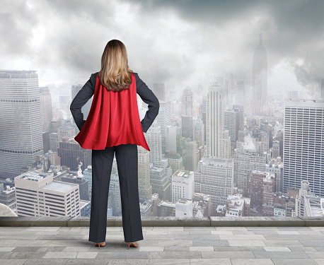 A rear view of a businesswoman wearing a red cape  looking out over the city below her. She is standing her hands on her hips as a gentle breeze flows through the cape.  A gray sky and patchy clouds hover over the city in the distance.