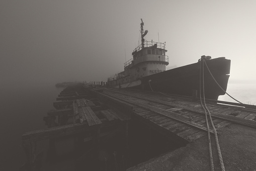 A foggy, night time view of an abandoned tugboat moored alongside a disused pier in heavy fog.  Long exposure, toned black and white.