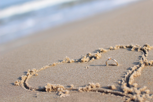 Engagement ring placed right in center of a heart drawn on a beach sand.