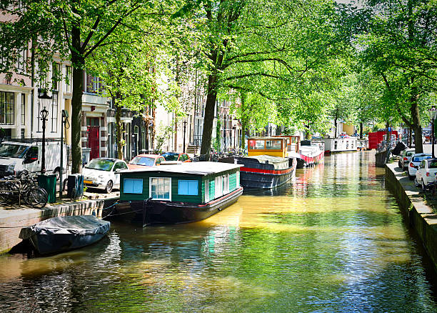 Canal of Amsterdam with green trees and boats anchored at the canal. Idyllic water street in Amsterdam, the Netherlands.