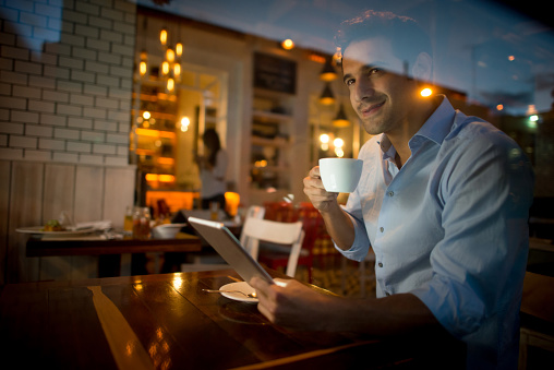 Handsome Latin American man reading at a cafe on a digital tablet while drinking a cup of coffee