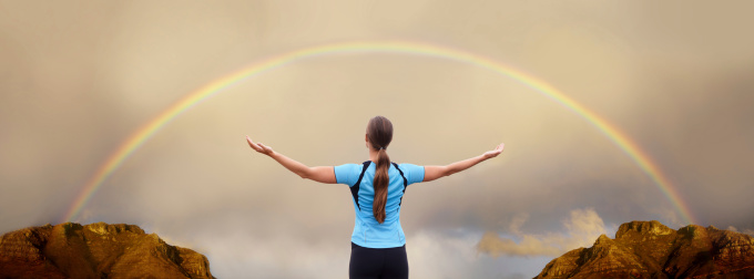 A young woman standing with arms outstretched in front of a rainbow
