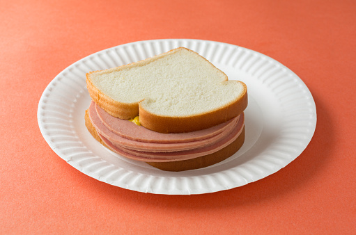 A several slices of bologna sandwich with mustard and white bread on a paper plate atop an orange table top.