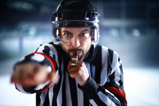 Male ice hockey referee blowing a whistle during a game and looking at camera.He's showing foul sign.Wearing full referee uniform and helmet.He's middle aged caucasian man.