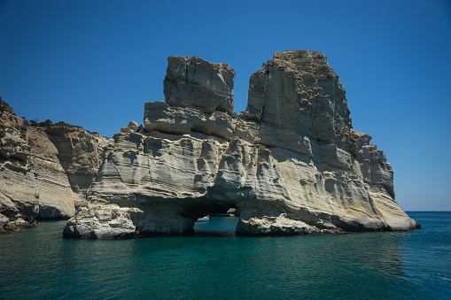 A serene view of the crystal-clear turquoise waters of the Blue lagoon of Comino island (Mediterranean Sea) in Malta. The water is surrounded by rocky cliffs, and a white boat can be seen in the distance. The sky is blue with fluffy clouds, and the vegetation is dry and sparse.