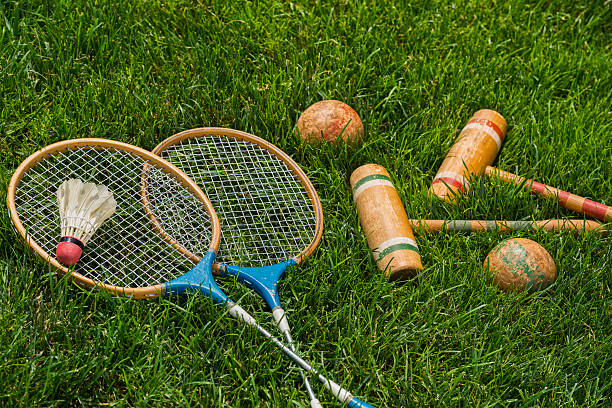 A pair of vintage wooden badminton rackets and a feathered shuttlecock lying in grass, with a pair of croquet mallets and balls. Great old fashion family fun games. 