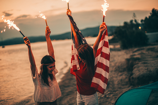 Young hipster girls at the beach, celebrating the fourth of July - by holding an American flag and fire torches up high 