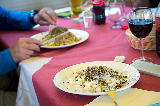 Senior tourist enjoying lunch, pasta(tagliatelle) with truffle on the top. on the summer terrace, Castelluccio, Umbria, Italy. There is a plate of gnocchi with truffle and red wine in front, out of focus. The man  is wearing a blue sweatshirt. Only his hands are visible. Nikon D800, full frame, XXXL. Higher ISO.