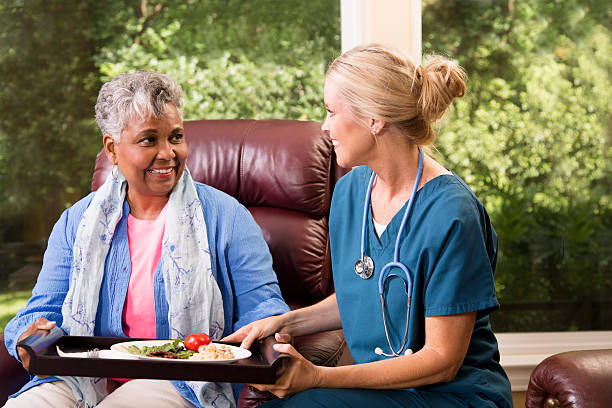 Home healthcare nurse with senior adult patient. Meal. Caring home healthcare nurse delivers a hot meal to an African descent senior adult woman at her home or nursing home facility.  The female patient is sitting in her comfortable chair while waiting on her dinner. Backyard can be seen through the windows.  meals on wheels photos stock pictures, royalty-free photos & images