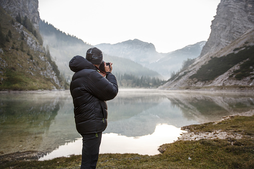 Photographer on assignment, holding a camera, taking photos of beautiful mountain landscape in the morning by a mountain lake with winter mist covered surface.