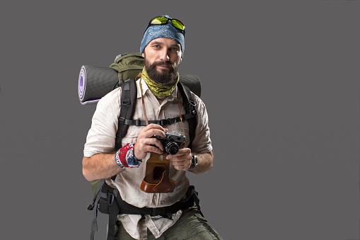 The tourist with camera. Portrait of a male fully equipped tourist with backpack. He photographing something on gray background, 