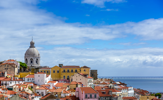DSLR picture of the City of Lisbon in Portugal on a nice summer day. Buildings with yellow and orange roofs and walls and a church are visible. The sky is blue with few clouds.