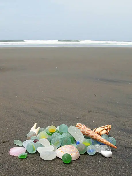 A pile of seaglass and some shells on a black sand beach, with the Pacific surf crashing in the background.