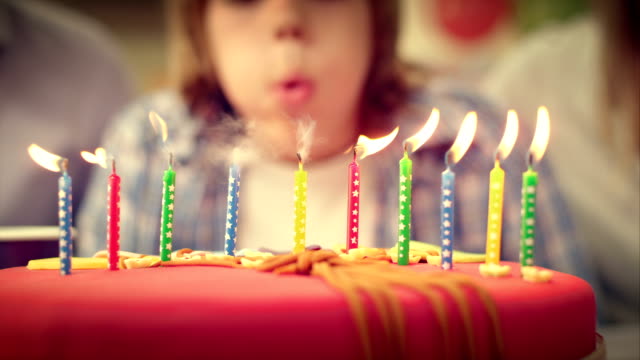 Slow motion shot of a nice birthday cake in the front with ten candles on it. The boy in the background leans over and blows the candles out. Video clip colour graded to achieve special look.