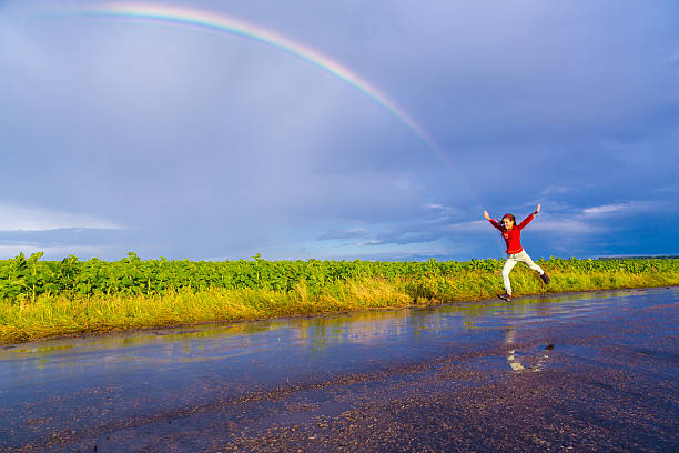 Happy girl jumping on wet road with rainbow after rain, with copy-space