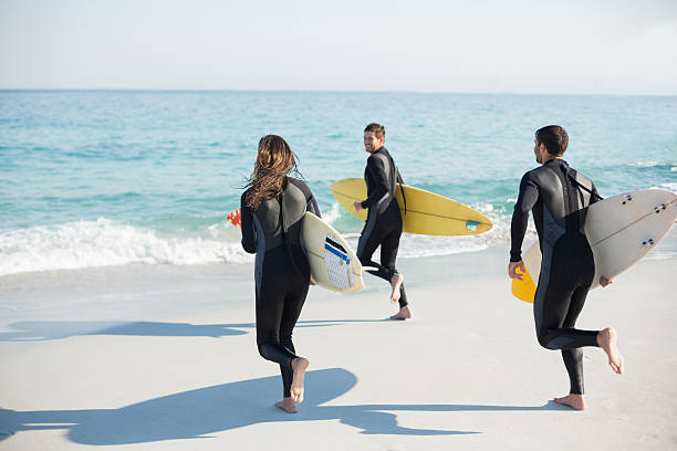 Group of friends on wetsuits with a surfboard Group of friends on wetsuits with a surfboard on a sunny day at the beach wetsuit stock pictures, royalty-free photos & images